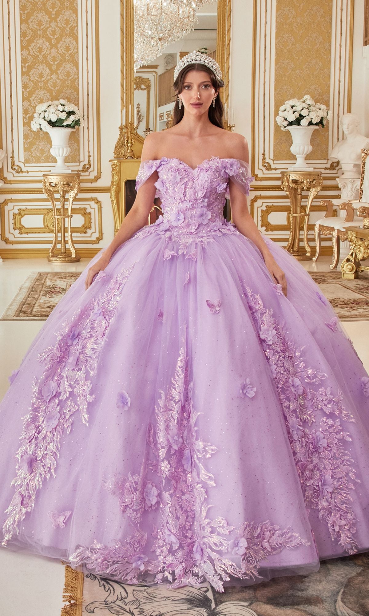 A woman stands in an ornate, gold-accented room wearing the Quinceanera Dress 15713 by Ladivine, a lavender ball gown with floral embroidery and butterfly appliques, her tiara enhancing the regal ambiance.